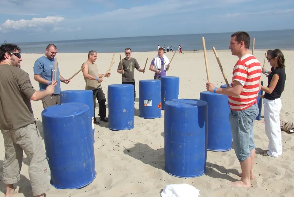 Uczestnicy bębnią pałkami w niebieskie beczki na plaży