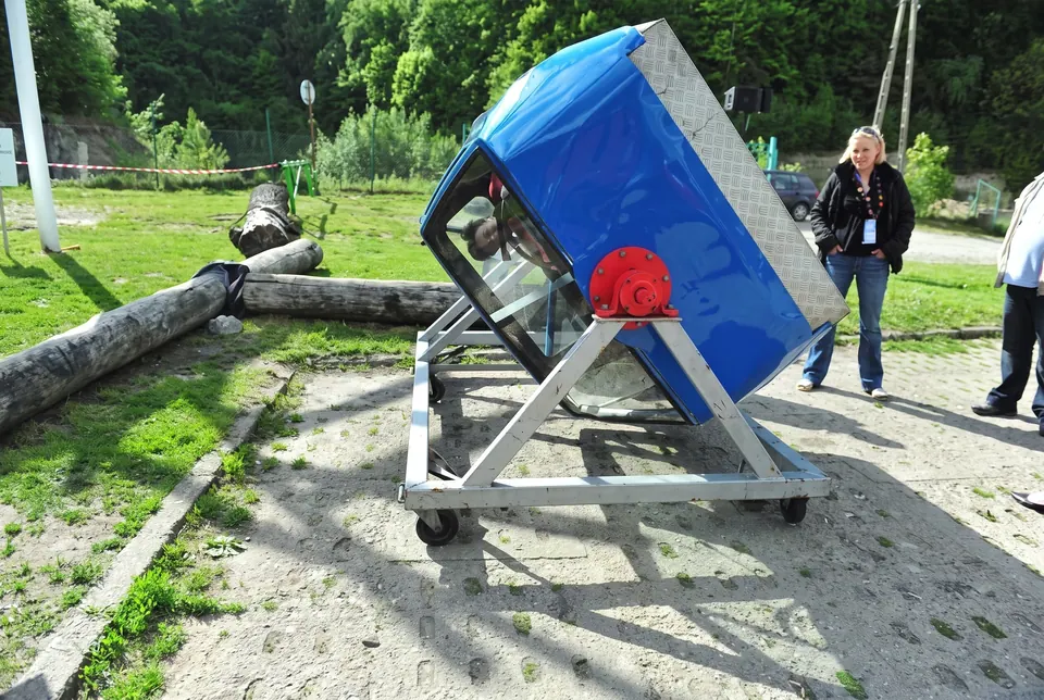 Niebieska kabina symulatora dachowania na stalowej ramie obok kłód