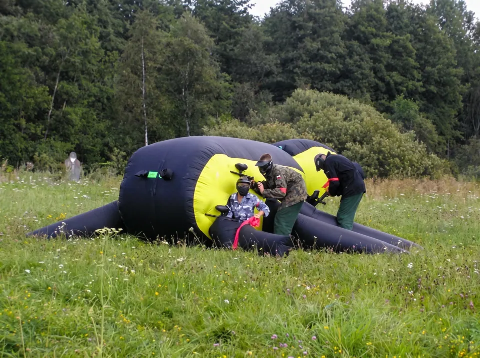 Gracze w maskach celują z markerów zza dmuchanej przeszkody na łące