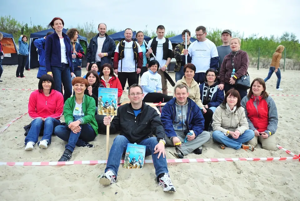 Grupa uczestników siedzi na piasku plaży w polu wyznaczonym taśmą