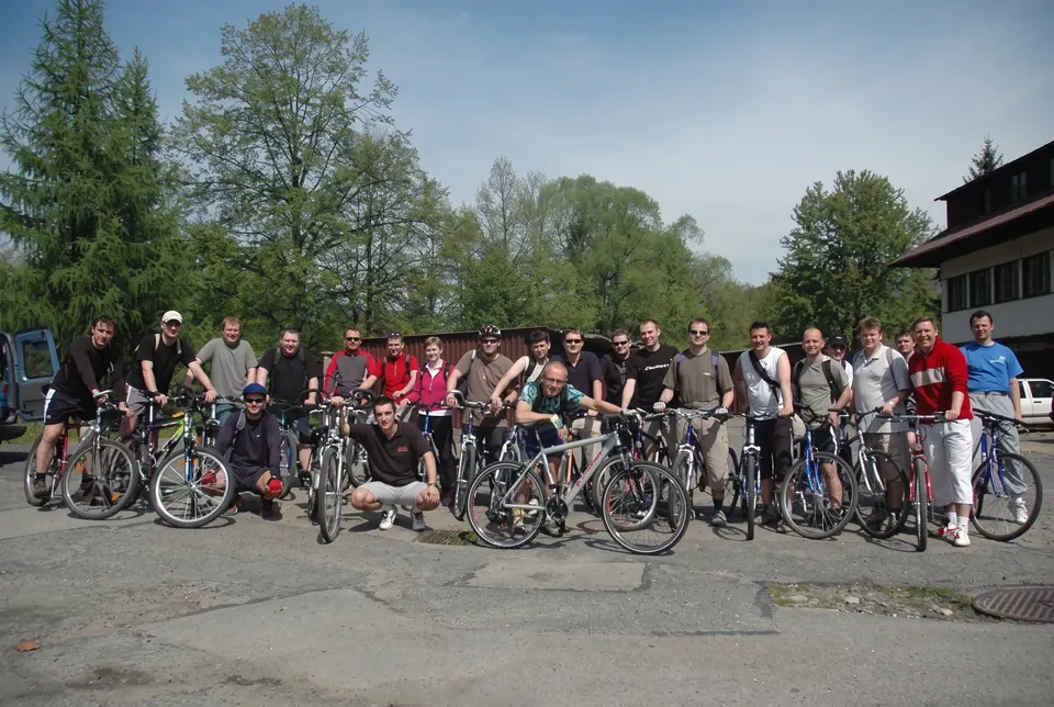 Grupa rowerzystów pozuje z rowerami na asfaltowym placu przy drzewach