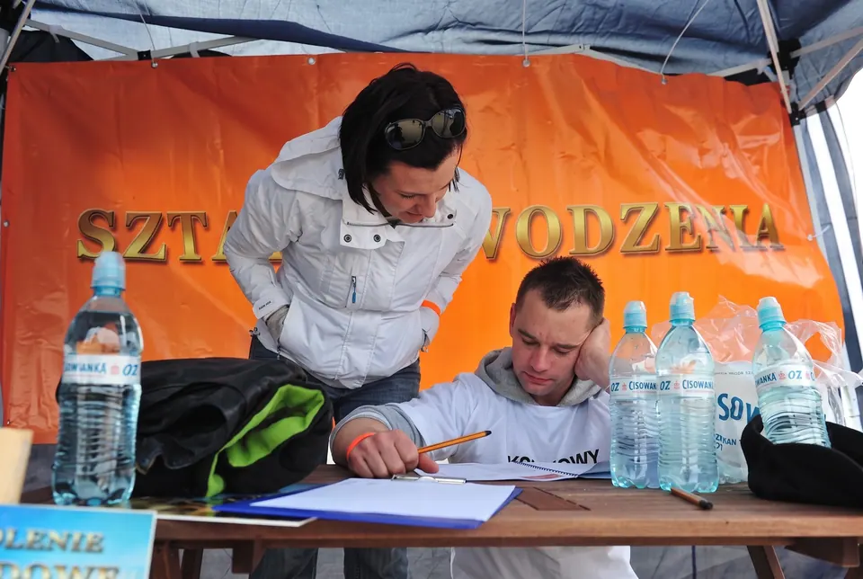 Uczestnicy zapisują wyniki przy stoliku z wodą pod pomarańczowym banerem