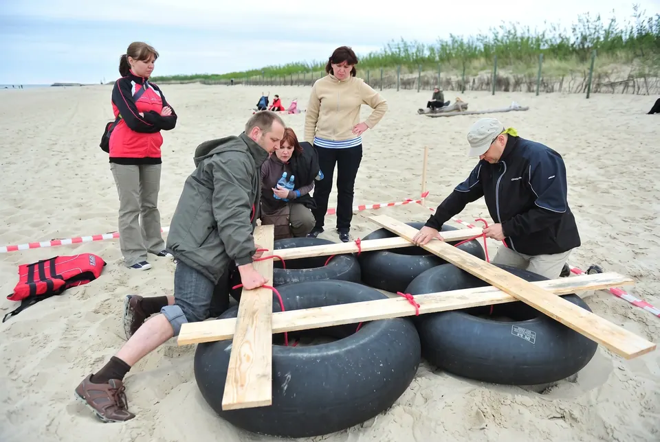 Uczestnicy budują tratwę z dętek i desek na piaszczystej plaży
