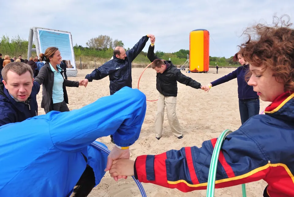 Uczestnicy trzymają się za ręce i przekładają hula-hoop na plaży