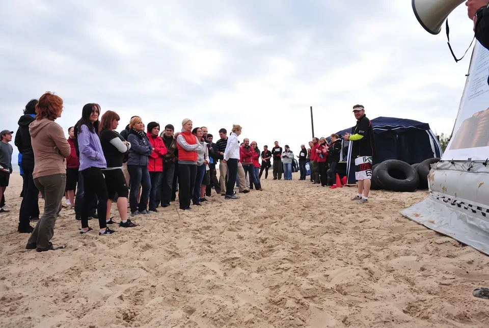 Długi szereg uczestników stoi na piaszczystej plaży przed konkurencją