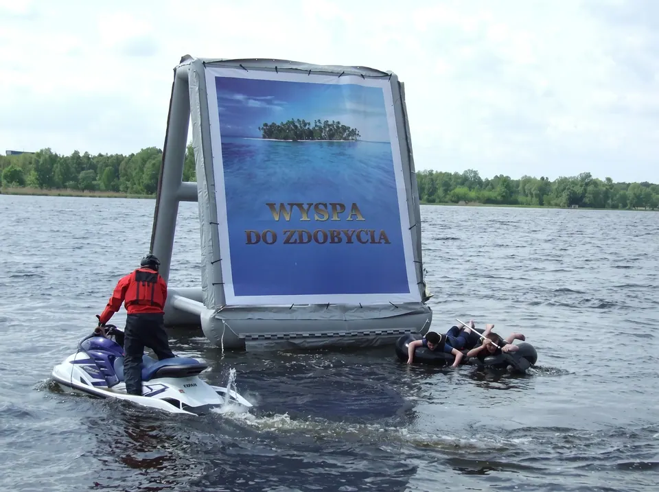 Uczestnicy na dętkach płyną do dmuchanej wyspy z plakatem na jeziorze